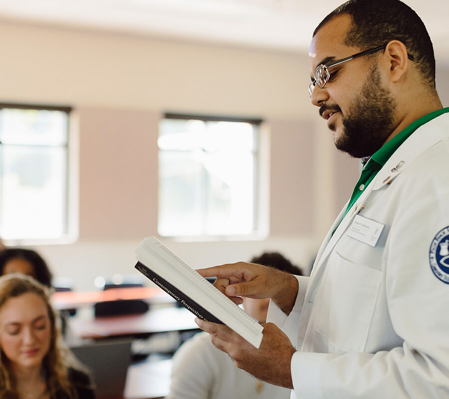 a male student standing in a classroom reading from a textbook
