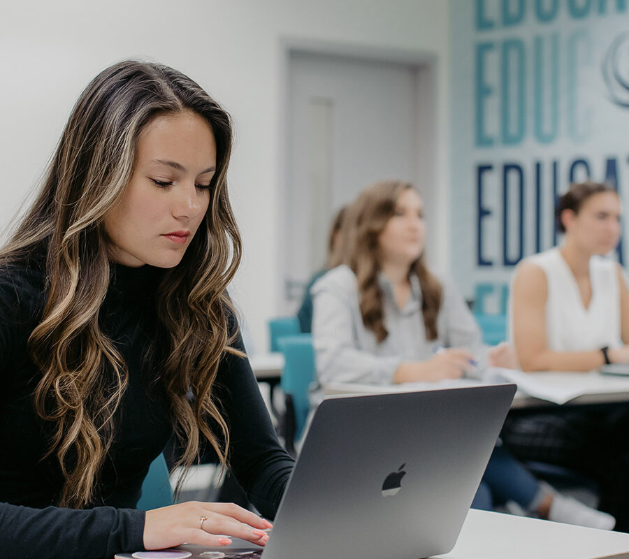 Students take notes on laptops during a lecture in a classroom.