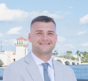 A smiling man in a light gray suit stands by a waterfront with a bridge and palm trees in the background under a blue sky.